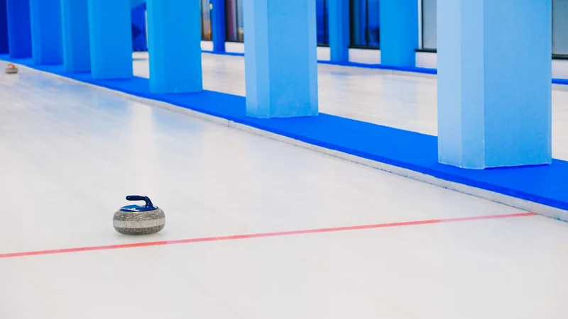 A curling stone resting on pale ice in an indoor rink with the sheet lines faintly visible in soft overhead light