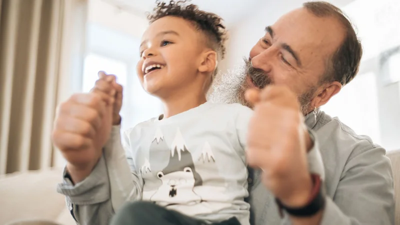 An older adult seated in a chair smiling while a young child stands beside them, both reaching their arms out in a playful pose