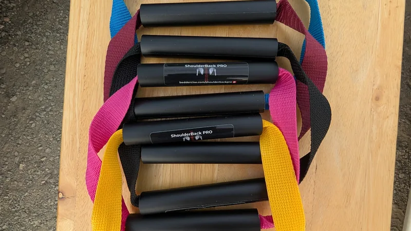 Five ShoulderBack Pro trainers with yellow, black, pink, burgundy and blue straps stacked on a wooden table