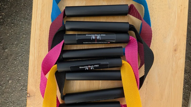 Five ShoulderBack Pro trainers with yellow, black, pink, burgundy and blue straps stacked on a wooden table