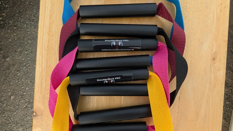 Five ShoulderBack Pro trainers with yellow, black, pink, burgundy and blue straps stacked on a wooden table