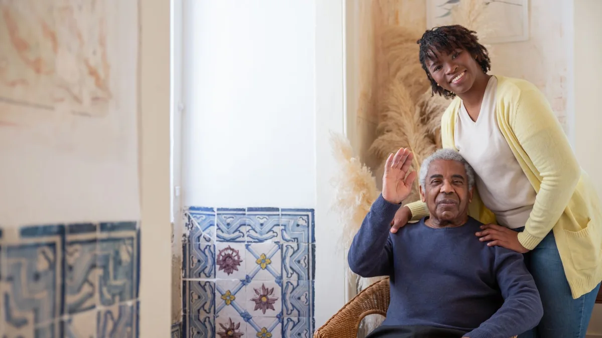 An adult child kneeling beside an older adult seated in a living room armchair, both relaxed, soft daylight from a window