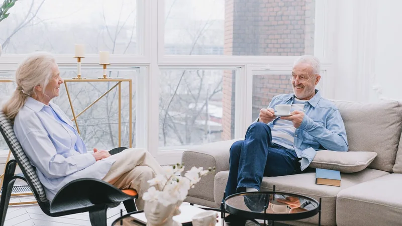 An older couple sitting side by side in armchairs by a window, smiling and talking