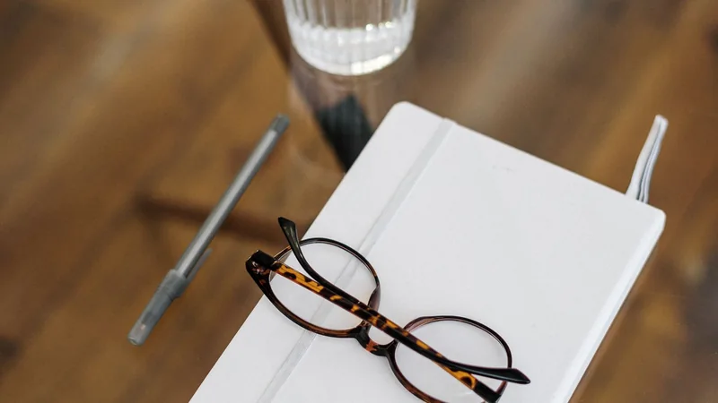 A glass of water on a kitchen table next to a notepad and a pair of reading glasses