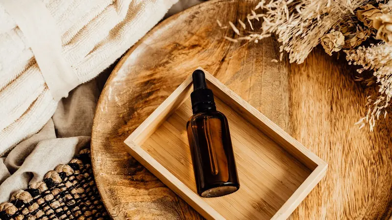 A small glass bottle of plain oil beside a folded towel on a wooden bench, with soft daylight from a window.