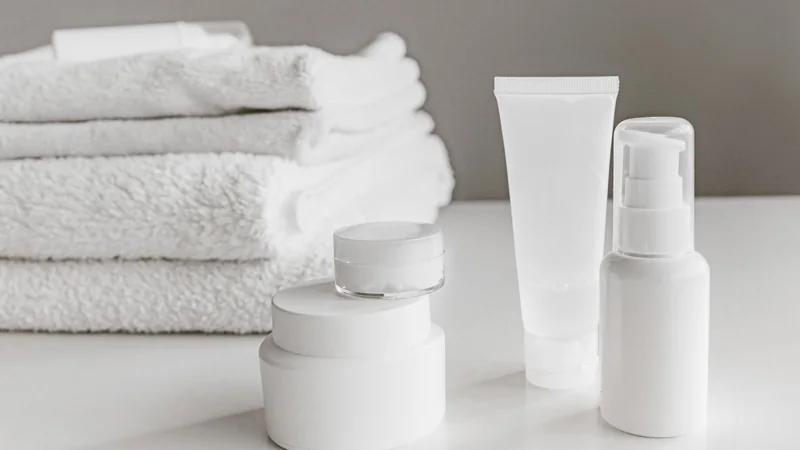 A small tube of cream on a bathroom shelf next to a folded towel and a glass of water.