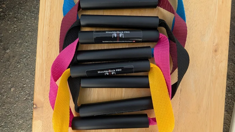 Five ShoulderBack Pro trainers with yellow, black, pink, burgundy and blue straps stacked on a wooden table