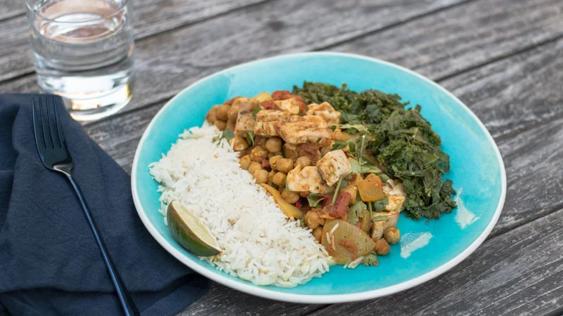 A bowl of lentils, chickpeas and tofu cubes on a wooden table next to a glass of water