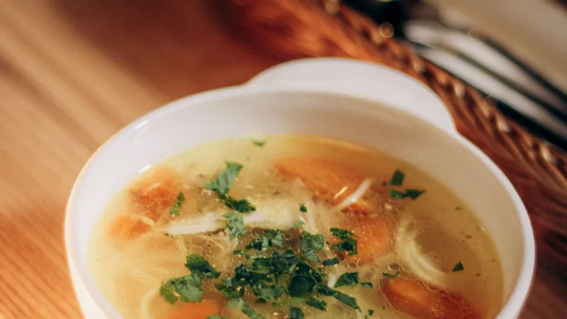 A bowl of clear broth on a wooden table beside a spoon and a sprig of herbs