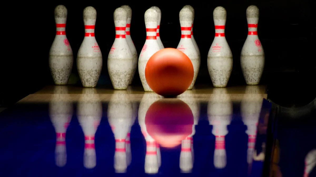 A bowling ball resting on a polished wooden lane near a row of white pins, in soft overhead light