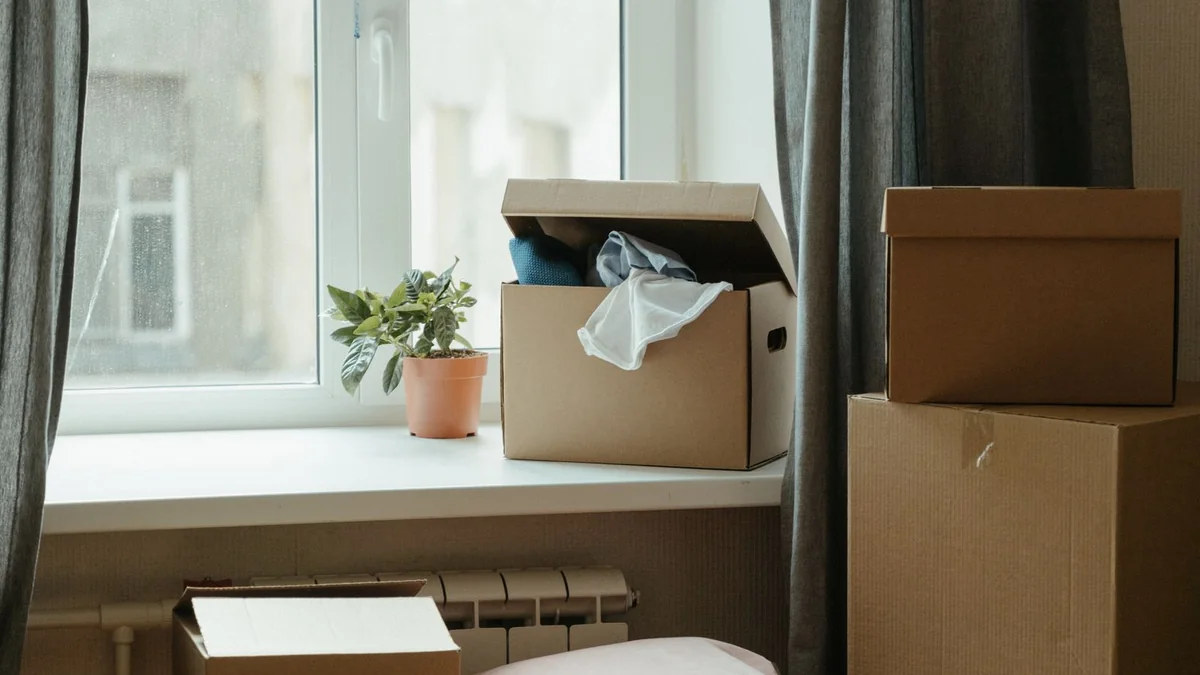 Stacked cardboard moving boxes beside a sunny front window in an empty living room