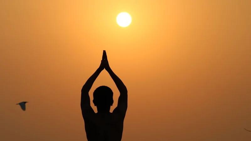 A person in a simple standing pose on a yoga mat outdoors in the early morning, arms raised overhead with the sun low on the horizon