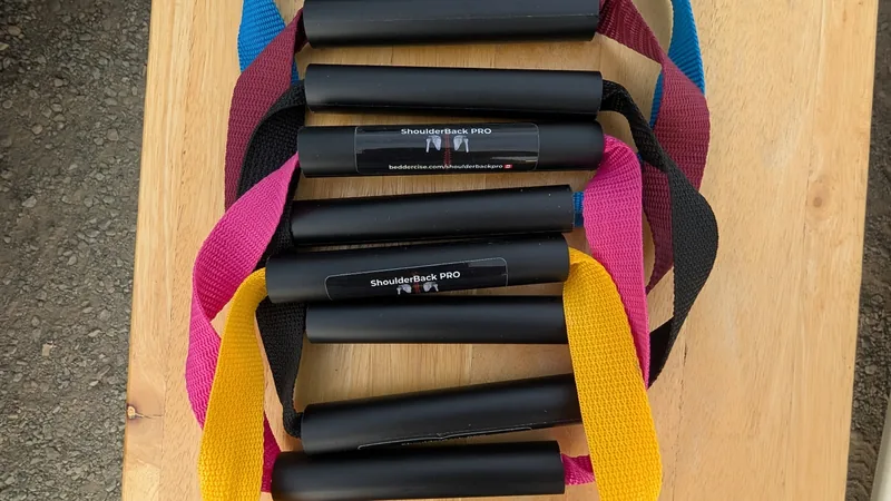 Five ShoulderBack Pro trainers with yellow, black, pink, burgundy and blue straps stacked on a wooden table