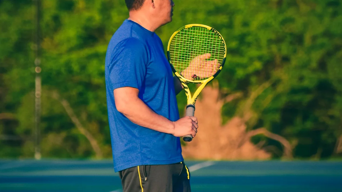 A person in a light shirt standing on a sunny sports court, mid-stride with a racket held at the side.